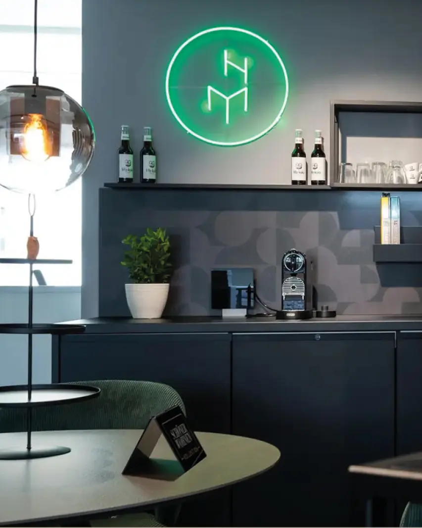Dunkle Küchenzeile mit Regal, Kaffeemaschine und Flaschen, grünes Neonlogo an der Wand, Tisch und Lampe im Vordergrund